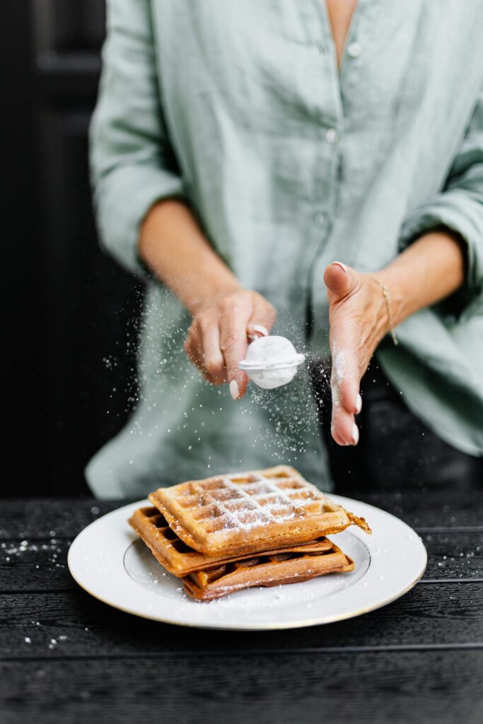 Close-up of a woman adding powdered sugar to freshly made waffles on a plate.